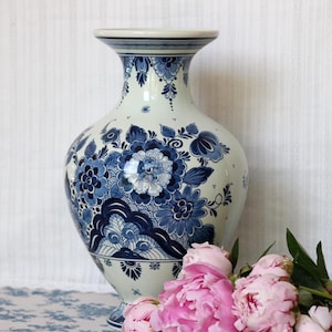 Può includere: Un vaso in ceramica con base bianca e complessi motivi floreali blu insieme a volute decorative. Il vaso presenta un bordo ampio e svasato, un corpo arrotondato e una base a piedistallo. Il design include petali e foglie dettagliate in blu.
