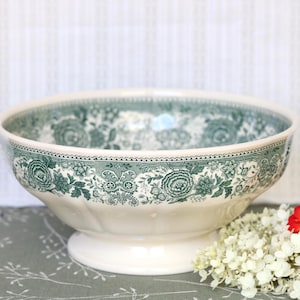 May include: A cream-coloured ceramic bowl featuring a dark green floral transferware pattern around the exterior rim. The bowl has a wide, circular opening, a rounded body, and a pedestal base. The interior is white with a matching green floral border.
