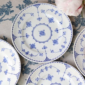 Może przedstawiać: Zestaw białych talerzy porcelanowych z niebieskim wzorem kwiatowym, centralnym motywem koła i żebrowanym brzegiem. Talerze są ułożone w grupę, prezentując szczegółowe niebieskie ilustracje botaniczne oraz delikatną, prążkowaną fakturę powierzchni.