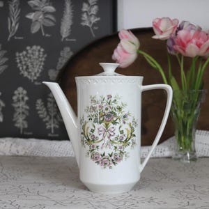 May include: White ceramic coffee pot with a floral design in pink, green, and purple. The pot has a long spout, a handle, and a lid. The background includes a vase of pink tulips and a botanical print.