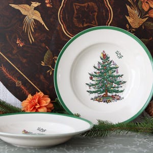 Może przedstawiać: Dwie białe ceramiczne miski z zielonym brzegiem, każda z centralną ilustracją udekorowanej choinki z kolorowymi ozdobami i prezentami, na ciemnobrązowym okrągłym tle ze złotymi motywami ptaków.