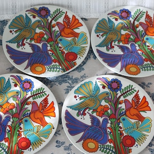 Puede incluir: Cuatro platos redondos de cerámica blanca con una vibrante ilustración repetida de aves estilizadas en tonos azul, naranja y dorado, posadas entre coloridas ramas florales. El diseño es consistente en los cuatro platos.