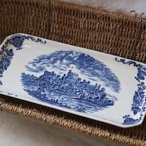 May include: A rectangular white ceramic serving tray featuring a blue transferware pattern. The centre depicts a detailed castle landscape, while the edges are decorated with ornate floral scrollwork and scalloped handles on both sides.