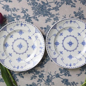 May include: Two white ceramic plates with blue floral designs. The plates have a central medallion and a patterned border. Purple tulips and blue flowers are arranged around the plates on a floral patterned tablecloth.