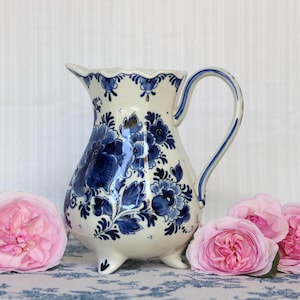 May include: A cream-colored ceramic pitcher featuring a scalloped rim, a curved handle, and three small feet. The surface is decorated with intricate dark blue floral patterns and foliage designs, complemented by three soft pink roses.
