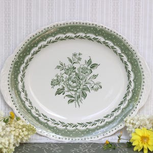 May include: An oval ceramic serving platter featuring a cream-colored center with a green floral bouquet illustration. The rim is decorated with a wide green border containing a repeating leaf pattern and a dotted texture with scalloped edges.