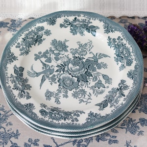 May include: A stack of three round ceramic plates featuring a cream-coloured base with a dark teal blue transferware pattern. The design includes intricate floral arrangements, leafy vines, and stylised birds centred on each plate.