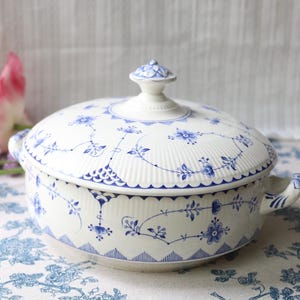 May include: A white porcelain tureen with a lid, featuring a delicate blue floral pattern and intricate line work. The round serving dish has two side handles and a small knob on the lid, with a ribbed texture along the top and sides.