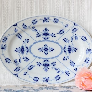 May include: An oval porcelain serving platter featuring a white base with a delicate blue floral pattern. The design includes a central oval medallion surrounded by symmetrical vine motifs and a ribbed edge texture along the outer rim.