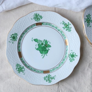 Peut inclure: Assiette en porcelaine blanche à bord festonné, ornée d'un motif floral vert et d'une bordure dorée. L'assiette a une surface texturée et un motif floral vert central. D'autres assiettes sont visibles en arrière-plan.