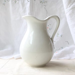 Op de afbeelding: Een witte keramische kan met een gebogen handvat en een tuit. De kan heeft een afgeronde body en staat op een lichtgekleurd oppervlak. De achtergrond heeft een witte doek met bloemenborduurwerk.