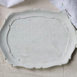 May include: A white, ornate ceramic tray with a floral pattern. The tray has a scalloped edge and decorative handles. The surface has a textured, embossed design. The tray is on a neutral-colored linen surface.