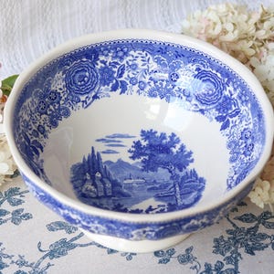 Może przedstawiać: Biała ceramiczna miska z niebieskim wzorem typu transferware. Wewnętrzna krawędź ozdobiona jest gęstym motywem kwiatowym przypominającym koronkę, a dno miski przedstawia szczegółowy krajobraz z budynkami, drzewami i górami w niebieskim kolorze.