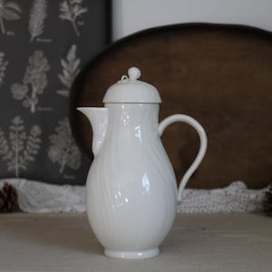 May include: A white ceramic coffee pot with a lid and handle. The pot has a textured, swirling design. The background includes a dark gray botanical print and a wooden tray. Pinecones and lace trim are also visible.