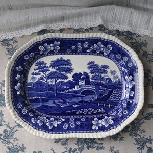 Op de afbeelding: Een ovale blauw-witte keramische serveerschaal met een landschapsscène. De schaal heeft een gedetailleerde illustratie van bomen, een brug en een gebouw. De rand is geschulpt en versierd met bloemmotieven.