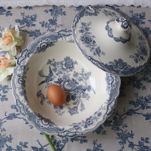 Può includere: Una ciotola da portata in ceramica color crema con coperchio abbinato, caratterizzata da un motivo blu con fiori e uccelli. La ciotola presenta un bordo smerlato e contiene un uovo marrone. Il design include dettagliate illustrazioni.