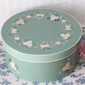 May include: A round, sage green storage box with a removable lid. The lid features a hand-painted wreath of pink flowers and green leaves. The side of the box is decorated with matching floral motifs. The box has a smooth, matte finish throughout.
