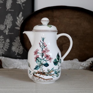 Könnte beinhalten: Eine weiße Kaffeekanne aus Keramik mit einer botanischen Illustration einer rosa blühenden Pflanze mit grünen Blättern und sichtbaren Wurzeln. Die Kanne hat einen passenden Deckel mit Knauf und einen dünnen braunen Rand am Deckel und Ausguss.