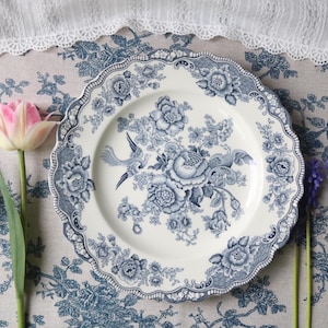 May include: A round ceramic plate with a scalloped edge featuring a blue and white floral pattern with a central bird illustration. The design includes detailed roses and foliage on a cream-coloured background, consistent with traditional transferware.
