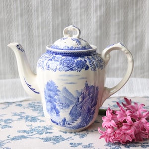 Op de afbeelding: Een vintage porseleinen theepot met een blauw en wit landschapsontwerp. De theepot heeft een gebogen tuit, een lusvormige handgreep en een deksel met een decoratieve knop. Het ontwerp bevat bloempatronen en een landschapsillustratie. Een tros roze bloemen staat in de buurt.