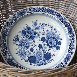 Puede incluir: Un plato de cerámica blanco con un diseño floral azul, colocado dentro de una cesta de mimbre. El plato presenta un grupo central de flores y un borde decorativo. La combinación de colores azul y blanco le da un aspecto clásico.