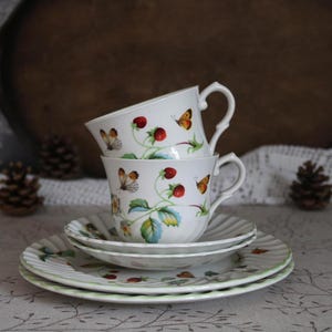 May include: Stacked white teacups and saucers with a strawberry and butterfly design. The teacups are stacked on top of each other, with the saucers below. The design includes red strawberries, green leaves, and colorful butterflies.
