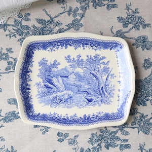 Op de afbeelding: Een rechthoekige keramische serveerschaal met geschulpte randen, voorzien van een crèmekleurige basis en een blauw transferpatroon. Het midden toont een landschap met bomen en een beek, omlijst door een decoratieve bloemenrand.