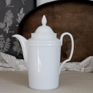 May include: A white porcelain coffee pot featuring a tall, cylindrical body, a curved handle, a pointed spout, and a matching lid with a finial. The upper section of the pot is decorated with a subtle, raised beaded texture around the rim.