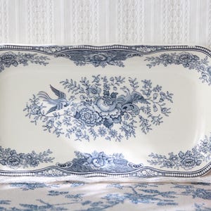 Pode incluir: Uma bandeja de servir oblonga de cerâmica com base na cor creme e um padrão de transferência azul escuro. O design apresenta um buquê floral central com pássaros, rodeado por uma borda decorativa de flores, folhas e motivos de frutas.