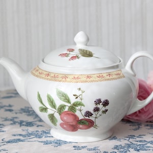 Peut inclure: Une théière en céramique blanche ornée d'une illustration botanique de prunes rouges et de mûres sombres sur le corps, avec un couvercle assorti décoré de petites baies rouges et de feuilles vertes, et une bordure décorative jaune et rouge.