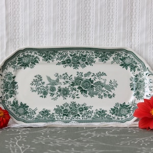 May include: An elongated, scalloped-edge ceramic serving platter featuring a white base with a detailed dark green transferware pattern of floral arrangements and birds. The rim is decorated with a repeating botanical border design.