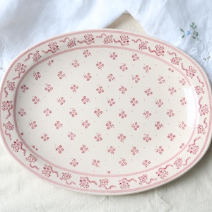 Può includere: Un piatto da portata ovale in ceramica con base color crema e un motivo ripetuto di piccoli fiori rossi. Il bordo presenta una decorazione con grappoli di fiori rossi e linee sottili, creando un design coerente su tutta la superficie.