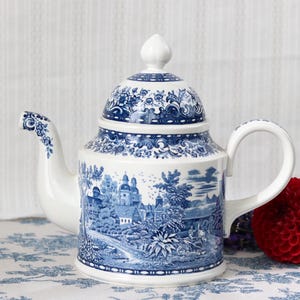Könnte beinhalten: Eine weiße Keramik-Teekanne mit einem blauen Transferdruck-Muster, das eine detaillierte Landschaft mit Gebäuden, Bäumen und Blattwerk zeigt. Der Deckel hat einen passenden blauen Blumenrand und einen spitz zulaufenden Knauf.