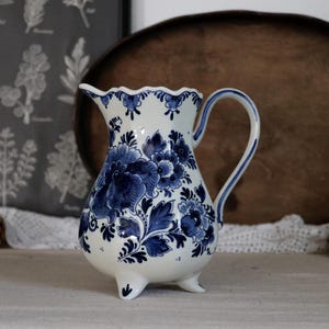 Può includere: Una brocca in ceramica bianca con un motivo floreale blu. La brocca ha un bordo smerlato, un manico curvo e tre piedini. Il motivo floreale blu copre il corpo, creando un aspetto classico e decorativo. La brocca è un oggetto decorativo.