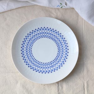 May include: White ceramic plate featuring a blue floral pattern in a circular arrangement. The plate has a textured surface and a wide rim. The blue pattern consists of small, stylised flower shapes. The plate is placed on a beige linen surface.