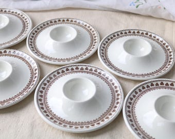 Może przedstawiać: Zestaw sześciu białych ceramicznych kieliszków do jajek, każdy z szeroką, płaską okrągłą podstawą i centralnie umieszczonym wgłębieniem. Krawędź każdej podstawy ozdobiona jest powtarzającym się brązowym geometrycznym wzorem małych kwiatów.