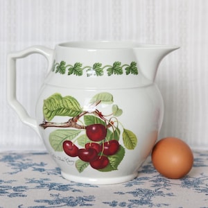 May include: A white ceramic pitcher featuring a botanical illustration of red cherries on a branch with green leaves. A green vine pattern encircles the upper rim. A single brown egg is positioned next to the base of the pitcher.