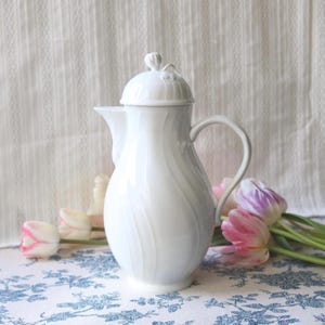 May include: A white ceramic coffee pot with a lid and handle. The pot has a curved design and a spout for pouring. The background includes pink and white tulips and a floral patterned tablecloth.