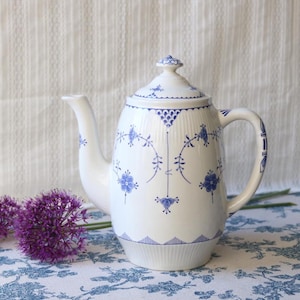 Peut inclure: Une théière en porcelaine blanche ornée d'un délicat motif floral bleu, de détails cannelés verticaux et d'un couvercle assorti. La théière présente une anse incurvée et un long bec, avec des motifs décoratifs bleus entourant le corps.