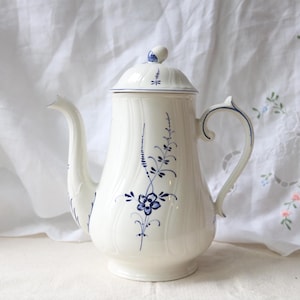 May include: A white ceramic coffee pot with a lid, handle, and spout. The pot is decorated with blue floral and vine patterns. The handle and spout are curved. The lid has a small knob on top. The pot is on a white surface.