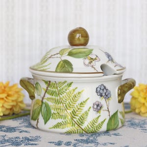 Puede incluir: Una olla de cerámica blanca con tapa y dos asas verdes, decorada con un patrón botánico pintado a mano de hojas de helecho verdes, pequeñas flores blancas y racimos de moras de color púrpura oscuro. La tapa tiene un pequeño pomo verde.
