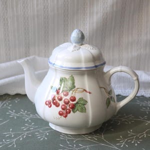 Può includere: Una teiera in ceramica bianca con corpo scanalato e coperchio abbinato con pomello azzurro. La teiera è decorata con un'illustrazione botanica di ribes rossi e foglie verdi, accentuata da una sottile linea blu lungo il bordo del coperchio.