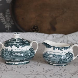 Może przedstawiać: Zestaw trzech ceramicznych kubków z nakrapianym, matowym wykończeniem. Kubki są w odcieniach kremu, jasnoszarym i ciemnoszarym. Każdy kubek ma prosty, minimalistyczny design, idealny do kawy lub herbaty.