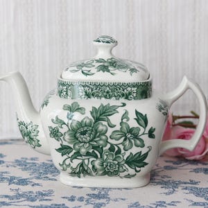 Może przedstawiać: Biały ceramiczny czajniczek ze szczegółowym zielonym wzorem kwiatowym i pasującą pokrywką. Czajniczek ma kwadratowy korpus z zaokrąglonymi rogami, zakrzywioną wylewkę i uchwyt. Zielony wzór zawiera misternie wykonane liście i kwiaty.