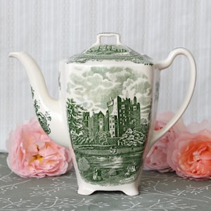 Pode incluir: Um bule de cerâmica cor de creme com uma estampa verde de transferência do Castelo de Blarney. O design retrata um castelo de pedra, árvores e figuras perto de um corpo de água. O bule tem um corpo quadrado, uma alça curva e uma tampa.