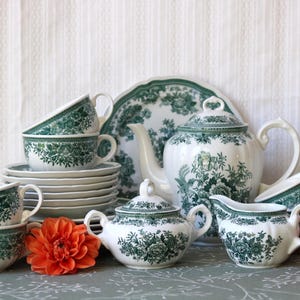 Könnte beinhalten: Ein weißes Keramik-Teeservice mit einem detaillierten grünen Blumenmuster. Das Set besteht aus einer Teekanne, einer Zuckerdose mit Deckel, einem Milchkännchen, mehreren Tassen und Untertassen. Die Teile sind mit botanischen Motiven in Dunkelgrün.