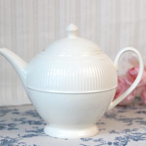 May include: A white ceramic teapot featuring a rounded body with vertical ribbed detailing on the upper section, a smooth lower base, a curved handle, a pointed spout, and a matching lid with a small knob handle on top.
