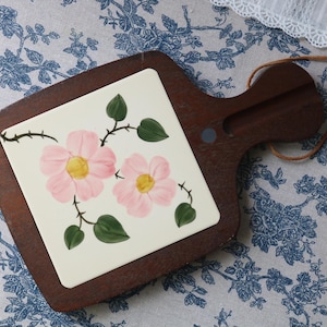 May include: A dark brown wooden serving board with a square ceramic tile inlay featuring a hand-painted design of two light pink flowers with yellow centers and green leaves on thorny stems, complete with a leather hanging cord attached to the handle.
