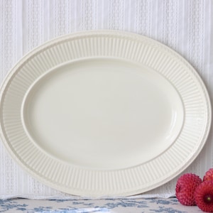 Pode incluir: Uma travessa de servir oval em cerâmica de cor creme, com uma base central lisa e uma borda larga decorada com um padrão de nervuras verticais repetitivas. A travessa possui um acabamento brilhante e um design simples e limpo.