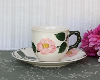 May include: A cream-colored ceramic teacup and saucer set featuring a hand-painted pink flower with a yellow center and dark green leaves. The teacup has a uniquely shaped, dark green handle with a textured, branch-like appearance.